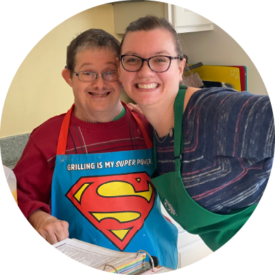 Two people wearing aprons lean together and smile in a kitchen
