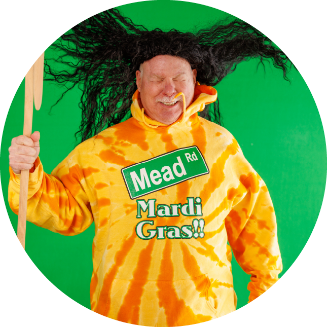 A person stands in front of a green screen and scrunches their face as wind blows their wig